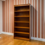 Load image into Gallery viewer, A tall wooden bookcase with five shelves stands against a wall with vertical pink and white stripes, set on a light wood floor.
