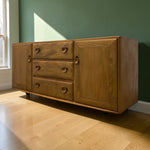 Load image into Gallery viewer, A mid-century modern wooden sideboard with three drawers and two cabinet doors, featuring round wooden knobs, set against a dark green wall and light wood flooring.
