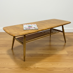 Load image into Gallery viewer, A mid-century modern wooden coffee table with a rounded rectangular top, angled legs, and a slatted lower shelf, shown on a wood floor against a white wall with a magazine on top.
