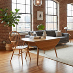 Load image into Gallery viewer, A mid-century modern wooden dining table with a drop-leaf style oval top and tapered legs, paired with a matching wooden spindle-back chair in a bright loft-style living room with brick walls, large windows, and a gray sofa.
