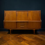 Load image into Gallery viewer, Mid-century modern wooden credenza with three upper cabinet doors, three lower drawers, and three side cabinets, shown against a dark blue wall and wood herringbone floor.
