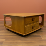 Load image into Gallery viewer, A mid-century modern wooden coffee table with two drawers, featuring a warm honey-wood finish, rounded edges, and circular wooden knobs, set against a terracotta wall and wood-look floor.
