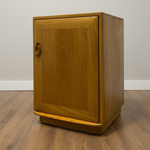 Load image into Gallery viewer, A mid-century modern wooden cabinet with a warm honey finish and a circular wooden pull handle, set on a wood floor against a plain wall.
