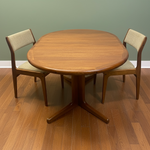 Load image into Gallery viewer, A mid-century modern wooden dining table set with two matching chairs, featuring a warm medium-brown wood finish and light beige upholstered seats, shown on a hardwood floor against a sage green wall.

