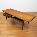 Load image into Gallery viewer, Mid-century modern wooden writing desk with a warm walnut finish, featuring a long rectangular top, tapered legs, and an open lower shelf with one drawer pulled out, set on a light wood floor against a white wall.
