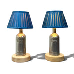 Load image into Gallery viewer, A pair of table lamps made from repurposed Absolut Vodka bottles, featuring blue pleated fabric shades, clear glass bodies, and round light wood bases, set against a plain white background.
