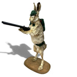 Load image into Gallery viewer, A taxidermy-style mounted rabbit figurine posed upright on a wooden base, wearing a green cap and backpack and aiming a black rifle, shown against a plain white background with a cast shadow.
