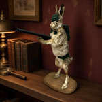 Load image into Gallery viewer, A taxidermy-style rabbit figure posed upright on a wooden surface, wearing dark green military-style clothing and holding a long gun. The scene is an interior room with a burgundy wall, books, a lamp, and framed artwork in the background.
