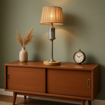 Load image into Gallery viewer, A mid-century modern table lamp with a pleated beige shade and brushed metal base sits on a wooden sideboard against a sage green wall, flanked by a vase with dried grass and a vintage alarm clock.
