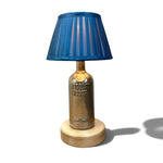 Load image into Gallery viewer, A table lamp featuring a blue pleated fabric shade, a vintage-style brass bottle base with 'Absinthe Vodka' text, and a round light wood footrest, shown on a white background with a soft shadow.
