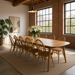Load image into Gallery viewer, A modern dining room setup featuring a light oak wood dining table and matching spindle-back wooden chairs, arranged on a textured beige area rug. The room has exposed brick walls, large black-framed industrial windows, a hanging pendant light, and a vase of white lilies on the table.
