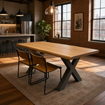 Load image into Gallery viewer, A modern live-edge wooden dining table with an X-shaped dark metal base, accompanied by two matching chairs with tan seats and black metal frames, set on a patterned area rug in a loft-style dining room.
