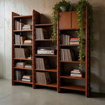 Load image into Gallery viewer, Three tall wooden bookshelves with a warm reddish-brown finish, styled with books, vinyl records, a turntable, and trailing green plants, set against a textured concrete wall and floor.
