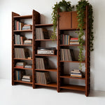 Load image into Gallery viewer, A set of three mid-century modern wooden bookshelves with a warm reddish-brown finish, styled with books, vinyl records, a turntable, and trailing green plants against a white wall.
