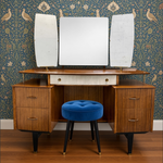 Load image into Gallery viewer, Mid-century modern wooden vanity table with three attached mirrors, a blue velvet stool, and brass hardware, set against blue patterned wallpaper and a wood floor.
