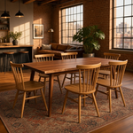 Load image into Gallery viewer, A warm wood dining table with six matching spindle-back chairs, styled in a modern loft dining room with brick walls, large windows, and a patterned area rug.

