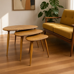 Load image into Gallery viewer, Three nested oval nesting tables in a warm medium wood finish, styled in a modern living room next to a yellow sofa and a potted green plant.
