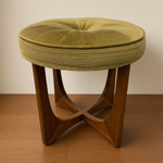 Load image into Gallery viewer, A round tufted ottoman stool with a textured olive green fabric cushion and a sculptural wooden four-leg base, shown on a wooden floor against a plain beige wall.
