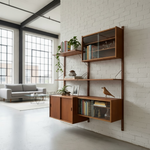 Load image into Gallery viewer, A mid-century modern floating wooden bookshelf and cabinet unit mounted on a white brick wall in a bright loft-style room with large windows and a gray sofa in the background.
